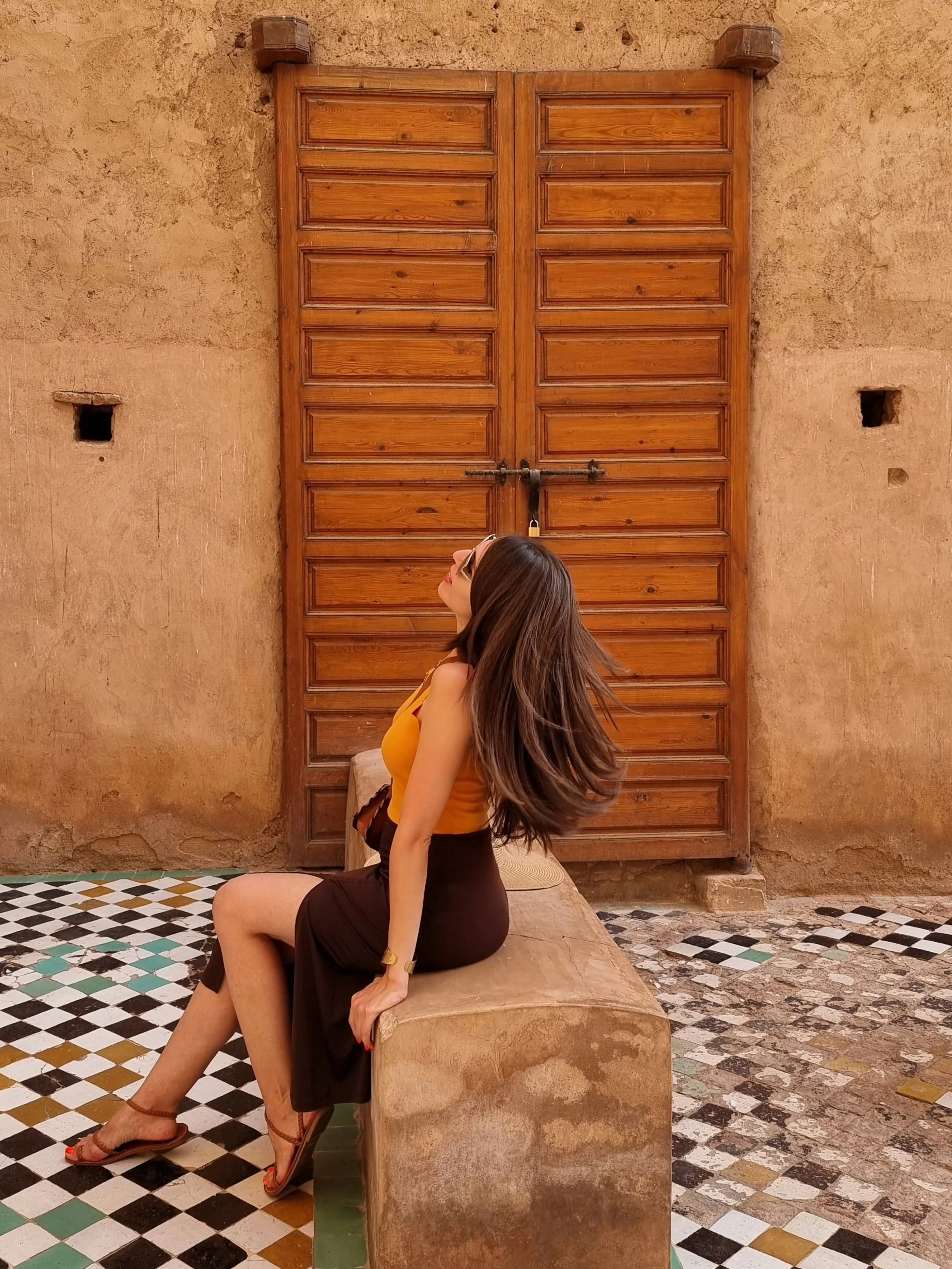 Deena leaning against a stone bench beside a carved wooden door.