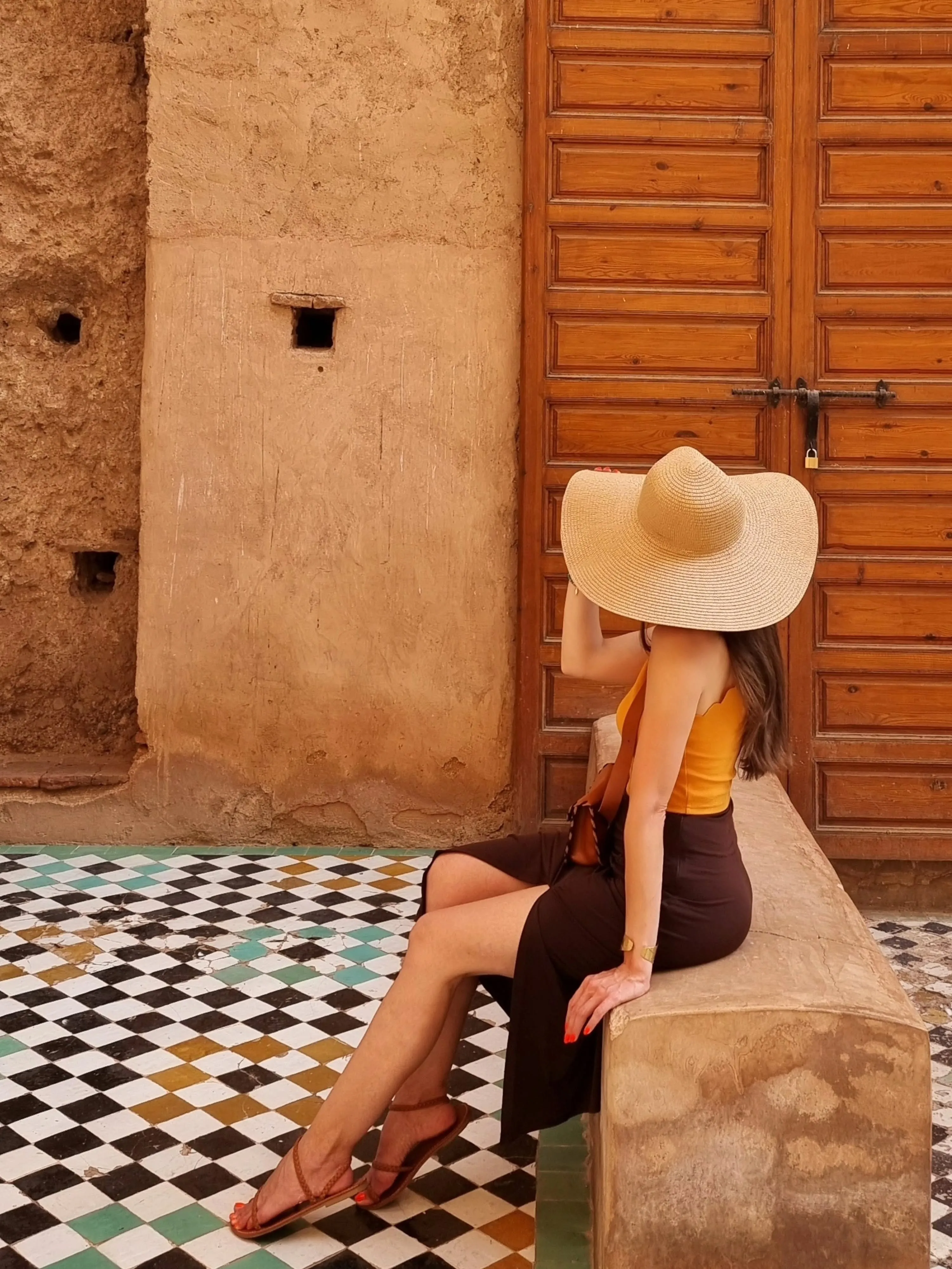 Deena sitting against an ochre wall in Andalusia, wearing a straw hat.