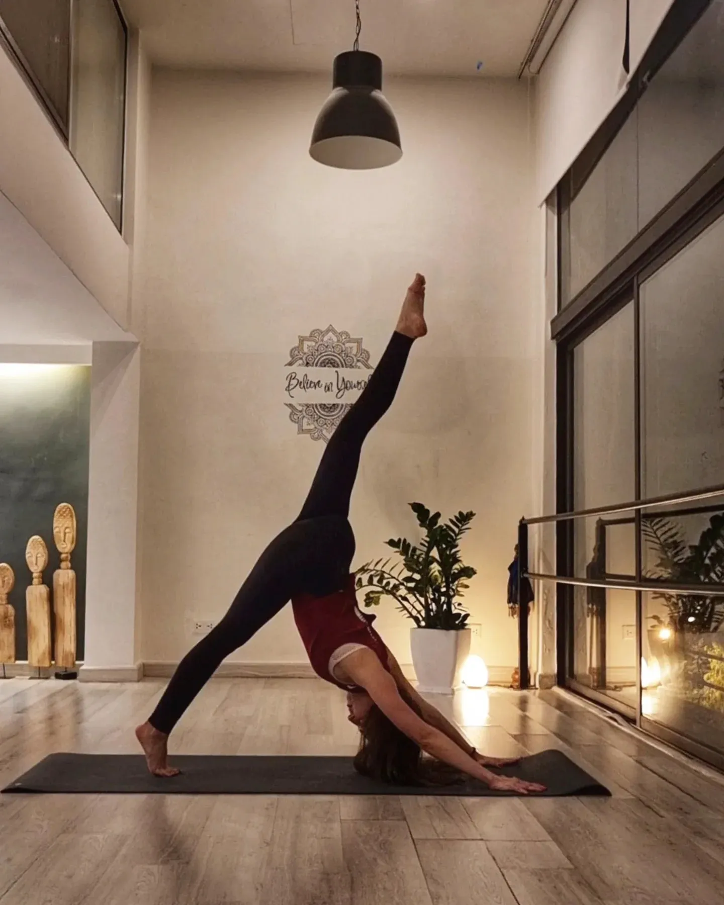 Deena in a standing split in a tall studio with warm lamps and daylight from a high window.